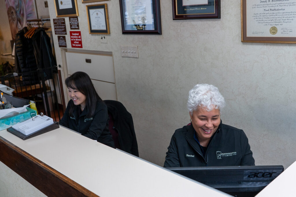 Two women sit at a reception desk, both smiling while working on computers in an office setting.